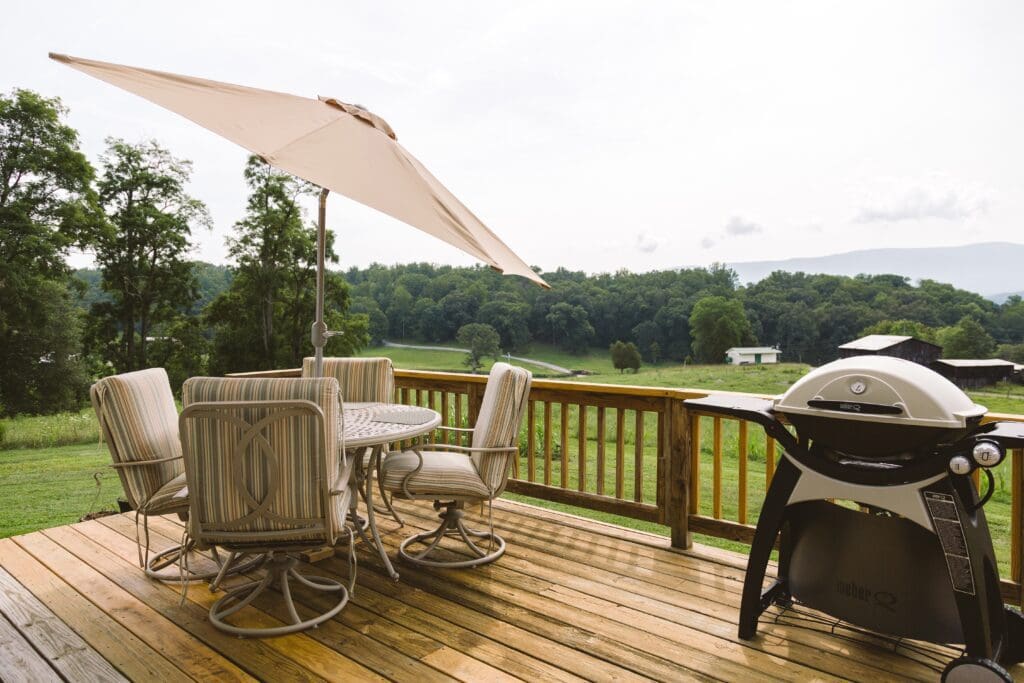 Outdoor furniture with table and umbrella and a grill on a deck with a wooden area in the background